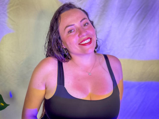 HoneyHotX met nat bruin haar, rode lippenstift, een zwart tanktopje, waarbij haar grote borsten zichtbaar zijn, en ze glimlacht. Ze draagt een sterrenketting, voor een paars-blauwe achtergrond..