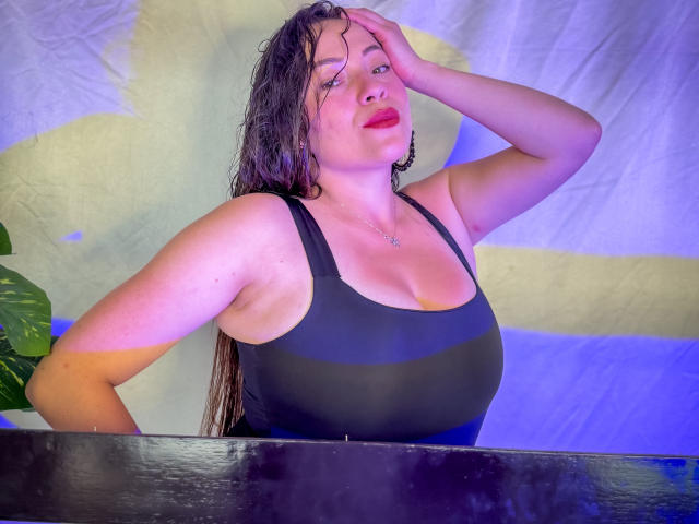 HoneyHotX con capelli castani umidi, rossetto rosso, canotta nera che mette in mostra il suo seno prosperoso, una mano sulla testa, in piedi vicino a una superficie scura, una pianta verde a sinistra..