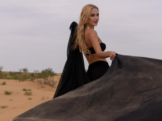 FloraDavis with long blonde hair, black strapless top, and black skirt, holding a black cloth, standing in a desert with sand and sparse greenery in the background.
