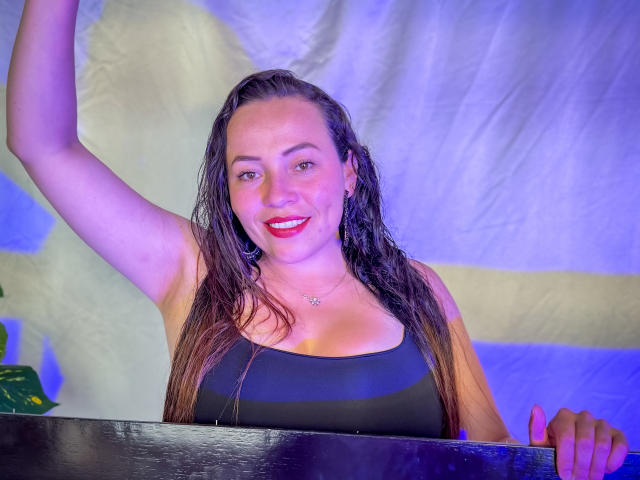 HoneyHotX met nat bruin haar, een zwart tanktopje, rode lipstick, lachend, arm omhoog, hand op een donker oppervlak, blauw-paars licht, een kleine sterrenketting, middelgrote borsten, aantrekkelijk, casual..
