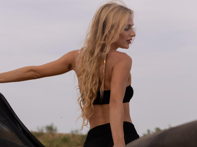 FloraDavis with long blonde hair, black strapless top, and black bottoms, showing her toned back and small ass, standing next to a black car, looking to the side.