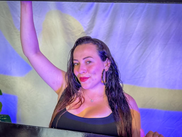 HoneyHotX con capelli castani umidi, grandi seni, canotta nera, orecchini a cerchio, e rossetto rosa, braccio alzato, sorridente, illuminazione blu e viola, leggero rossore sulle guance..