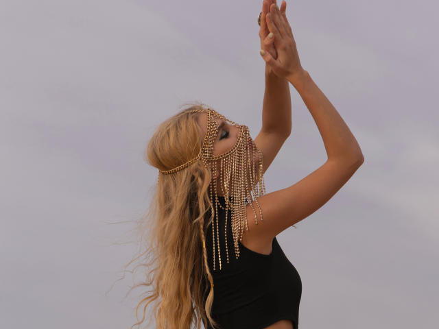 FloraDavis with wavy blonde hair, wearing a black crop top and gold chain headpiece with dangling beads, arms raised, eyes closed, against a cloudy sky.