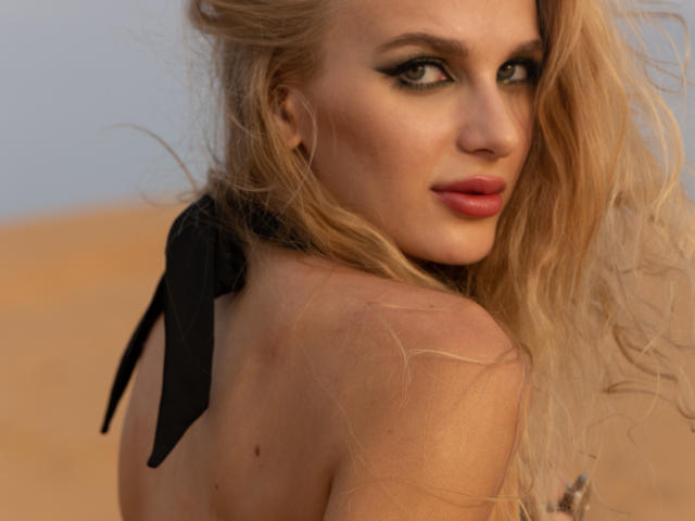 FloraDavis with long blonde hair, smoky eye makeup, and red lipstick, looking over her shoulder, wearing a black halter top, showing her bare back and small mole.