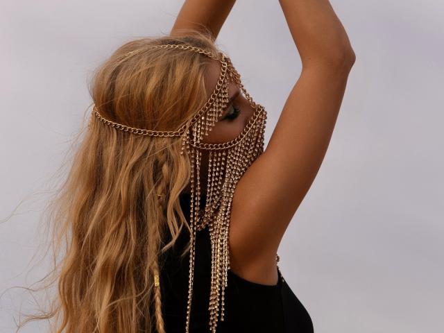 FloraDavis with long, wavy blonde hair, wearing a gold chain headpiece and matching chain mask, arms raised, black sleeveless top, tan skin, sexy, elegant, overcast sky.