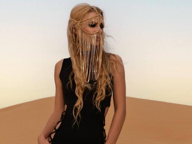 FloraDavis with long blonde hair, wearing a black dress and a gold chain mask covering her face, standing in front of a sandy dune. Her tits are visible.