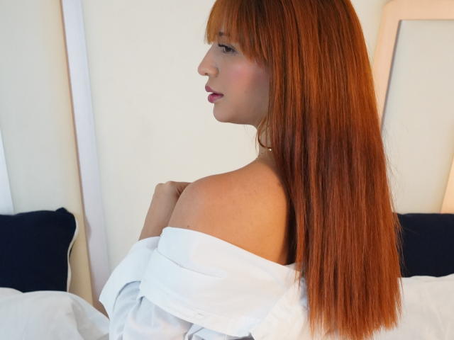 EmmaBonnet with long red hair, off-shoulder white shirt, looking side profile, showing bare shoulders and subtle cleavage, small pearl necklace, soft pink lipstick, white bed in background.