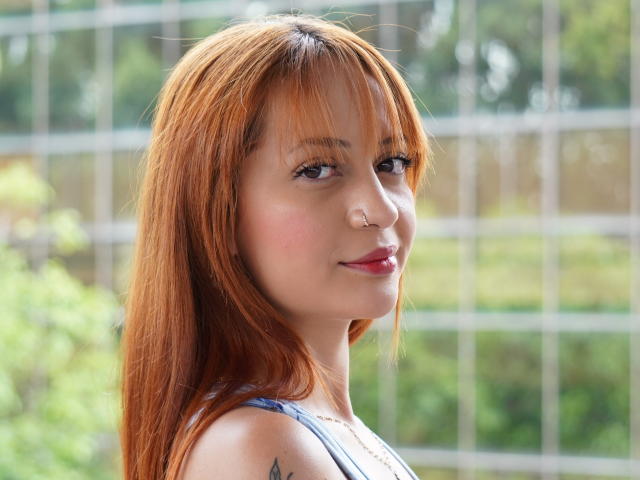 EmmaBonnet with red hair, nose ring, and tattoo on her arm, smiling at the camera, showing her tits in a sleeveless top. Her red lips match her hair.