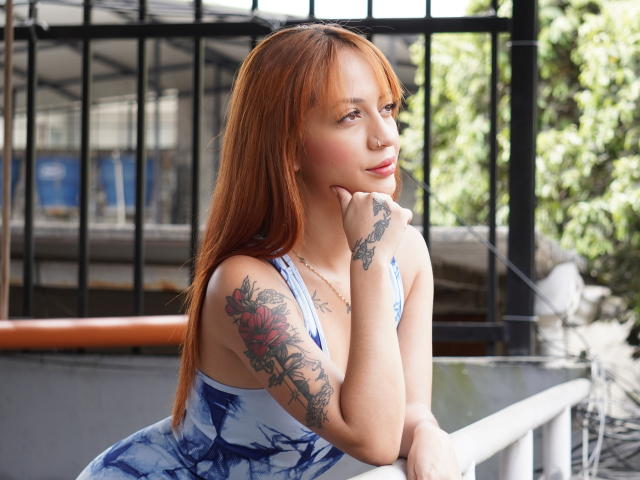 EmmaBonnet with red hair, tattoos on arm and side, blue tie-dye dress, resting chin on hand, looking side, soft smile, outdoor setting, black metal railing, sunlight.