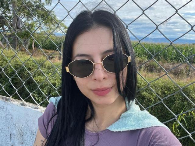 IsabellaJames with long black hair, wearing purple top, light blue hood, and gold aviator sunglasses, standing in front of a chain-link fence, showing a slight smile.