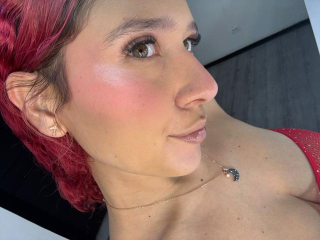 ValeryScot has pink-red hair, brown eyes, and a small nose ring. She wears a red sparkly top and a gold necklace with a blue butterfly pendant.