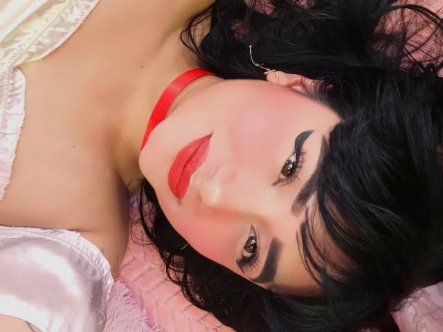 AnnieGrey tiene lápiz labial rojo, cabello ondulado negro y un collar rojo. Está acostada sobre una superficie rosa con mangas blancas, mirando hacia arriba con cejas oscuras y delineador de ojos..