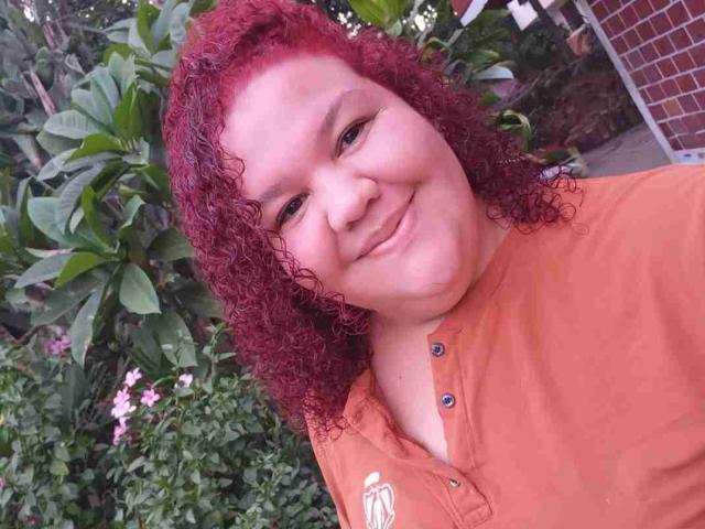 A TugorditaSexy tem cabelos ruivos cacheados, um rosto gordinho e um pequeno sorriso, vestindo uma camisa laranja, com folhas verdes e flores rosa ao fundo..