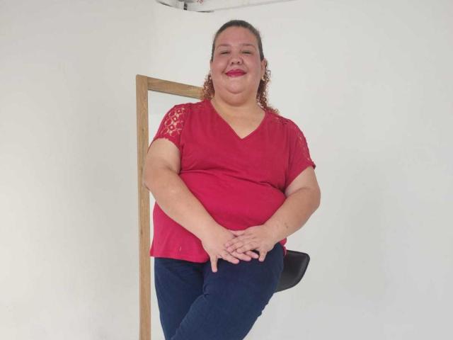 TugorditaSexy has a chubby face, curly brown hair, red V-neck shirt with lace, navy pants, and red lipstick, standing with hands on knees, smiling.