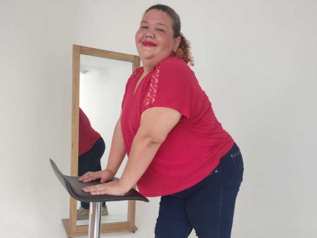 TugorditaSexy, une femme corpulente avec des cheveux bruns bouclés, un t-shirt rouge et un jean bleu, est appuyée sur un tabouret noir, souriant avec du rouge à lèvres, un miroir en arrière-plan..