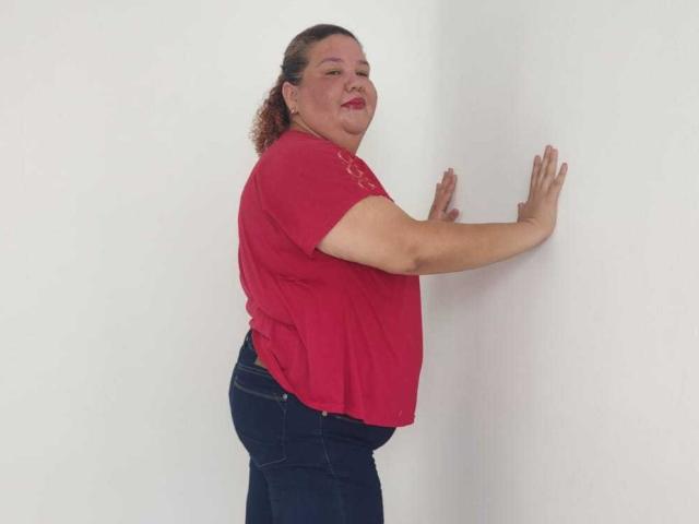 TugorditaSexy, a chubby woman with red shirt and blue jeans, stands against a white wall, smiling, hands raised, showing a slight belly and curly brown hair.