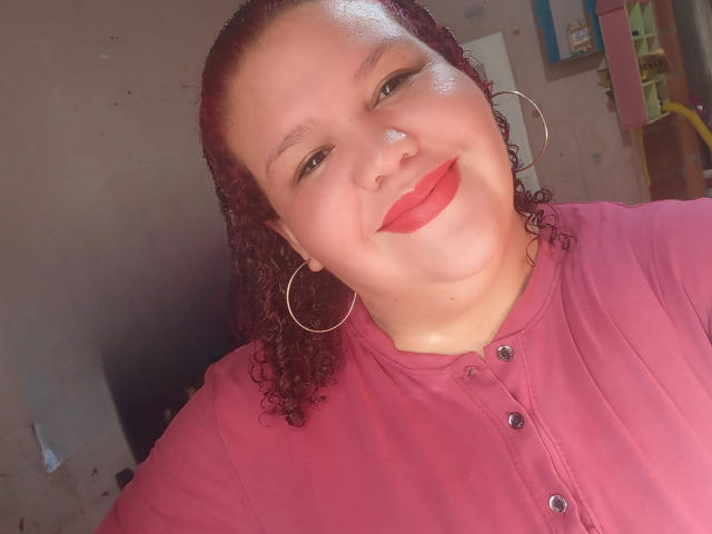TugorditaSexy with curly red hair, hoop earrings, and red lipstick, wearing a pink button-up shirt, smiling at the camera.