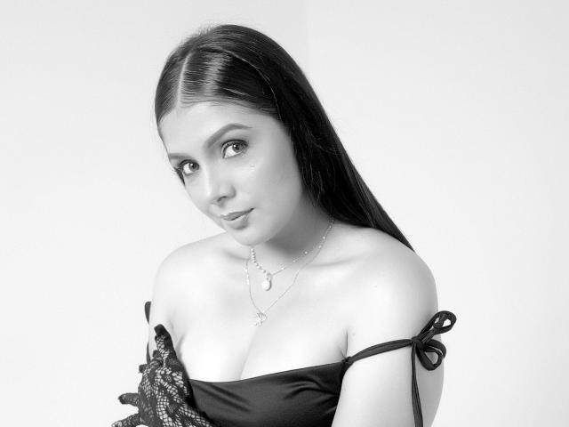DahianaLina has shiny skin, long dark hair, and is wearing a black off-shoulder top with lace. Her tits are visible, and she's looking at the camera with a slight smile.