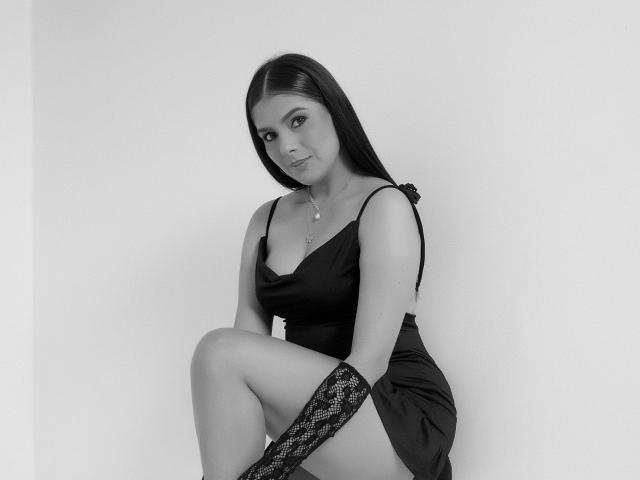 Black and white photo of DahianaLina with long dark hair, wearing a black satin dress, lace arm warmers, and a necklace, sitting with one leg up, smiling.