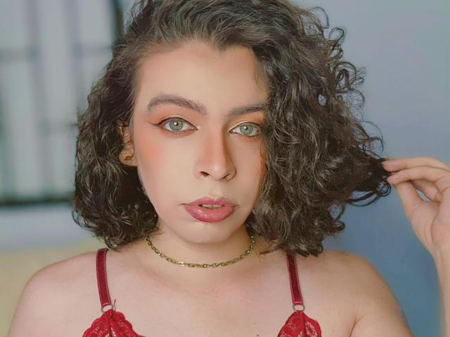 TsFoxCandy, met krullend bruin haar, draagt een rode kantbeha en een gouden ketting, heeft groene ogen, roze lippen en een lichte frons, en ze raakt haar haar aan met een serieuze uitdrukking..