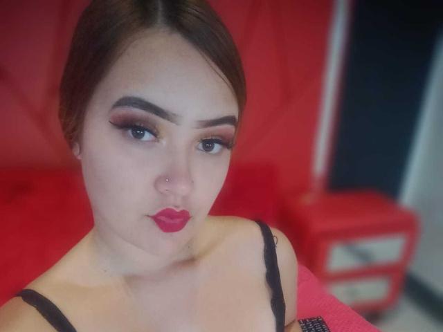 HencyKloss with dark brown hair, red lipstick, and black eyeliner, wearing a black strap, looking down, nose pierced, red background, and red furniture in the blurry background.