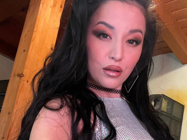 MissKataleya-hot with glossy pink lips, black wavy hair, orange eyeshadow, and a black choker, wearing a silver mesh top, looking at the camera with a sultry expression.