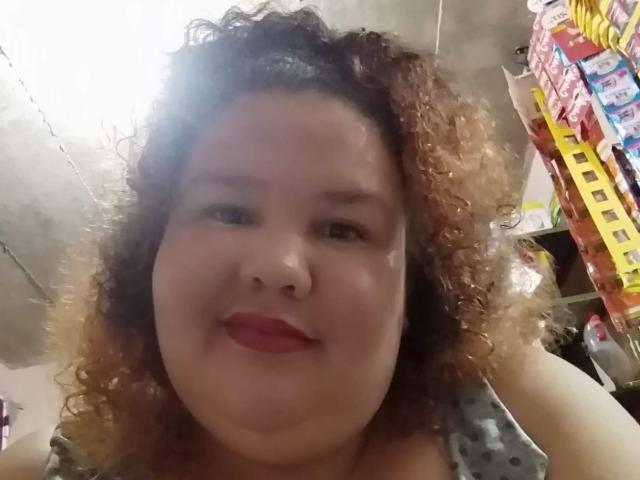 TugorditaSexy tiene el pelo castaño rizado, lápiz labial rojo y un rostro regordete con una ligera sonrisa, vistiendo una blusa de lunares, en un selfie con poca luz y ligeramente borroso..