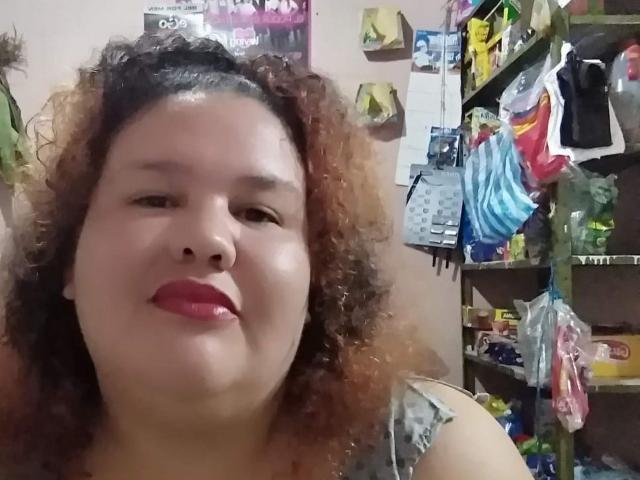 TugorditaSexy has curly brown hair, light skin, red lipstick, and is smiling slightly. She's wearing a gray top, and her boobs are partially visible. Shelves with clothes are in background.