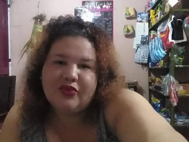 TugorditaSexy ha i capelli castano ricci, rossetto rosso e un viso paffuto con un'espressione accigliata, indossa una canotta grigia, in una stanza disordinata con scaffali..