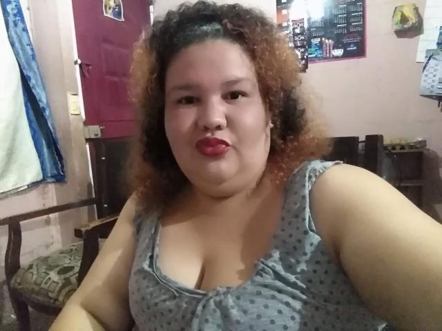 TugorditaSexy, uma mulher rechonchuda com cabelos castanhos cacheados, batom vermelho e uma blusa cinza com bolinhas, exibindo seus seios grandes e redondos, sentada em um quarto..