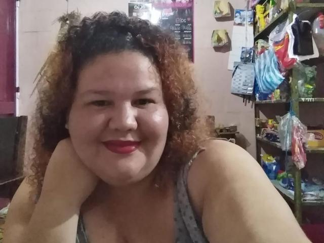 TugorditaSexy con capelli castani ricci, rossetto rosso, che sorride, indossa una maglietta grigia a pois, mostra leggermente il seno, appoggiata a un tavolo, in una stanza disordinata..