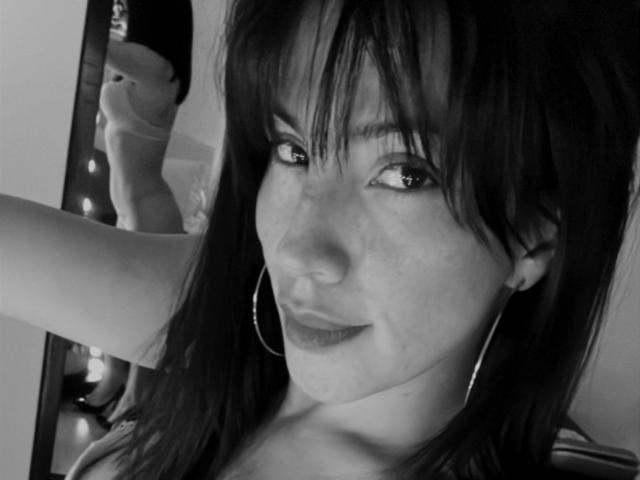 Black-and-white selfie of Liliysweet with medium-length dark hair, small hoop earrings, and a slight smile, showing a reflection of her bare ass in the mirror.