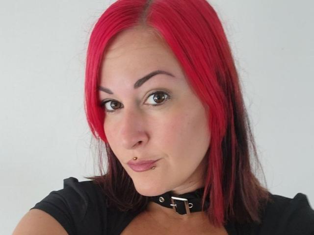 GennyRock has red hair, a black choker, a nose piercing, and a lip piercing. She's wearing a black shirt, looking at the camera with a slight smirk.