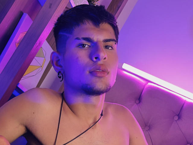 BradKiing, shirtless with short dark hair, has a black ear tattoo, slight goatee, and a necklace. Purple and blue lighting highlights his face and chest.