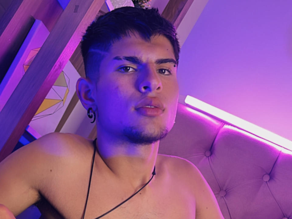 BradKiing, shirtless with short dark hair, has a black ear tattoo, slight goatee, and a necklace. Purple and blue lighting highlights his face and chest.