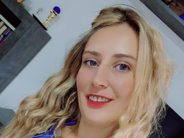 Adellee, mit blonden, gewellten Haaren, blauen Augen und rotem Lippenstift, lächelt in die Kamera. Ihr süßes Gesicht und ihr hübsches Lächeln werden hervorgehoben. Sie trägt ein blaues Oberteil..