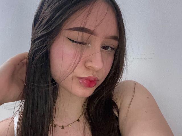 SophieHale with long brown hair, winking, pink lipstick, nose piercing, and a gold necklace, showing off her pouty ass-kissing face. Her right hand touches her hair.