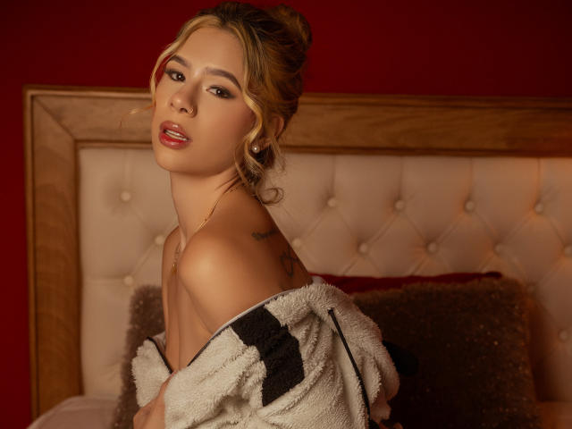 RileySwet with blonde hair in a bun, red lipstick, off-shoulder fluffy white jacket, small tattoos, looking over shoulder, dark red wall, wooden headboard, soft light.
