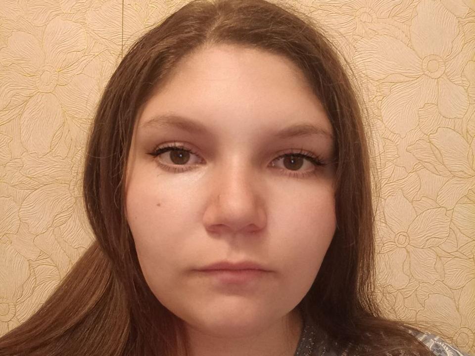 AdelinaRos hat braune Augen, langes braunes Haar und einen kleinen Schönheitsfleck auf ihrer rechten Wange. Sie schaut direkt in die Kamera mit einem neutralen Gesichtsausdruck..