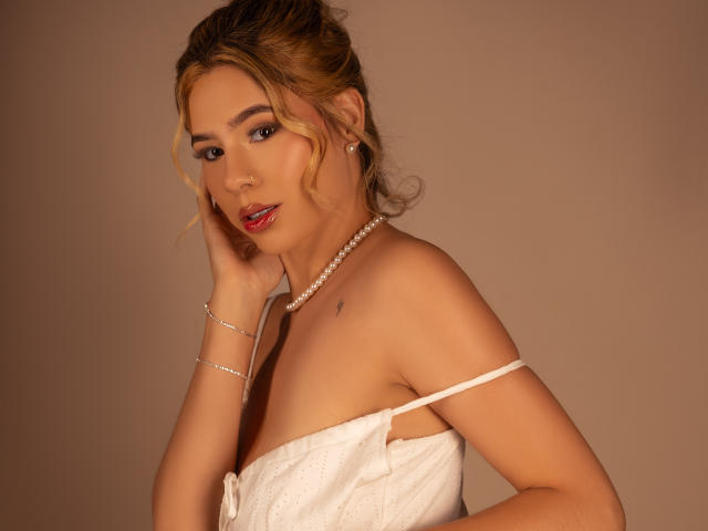 RileySwet with blonde hair, wearing a white strapless dress, pearl necklace, and silver bracelet, touching her cheek, looking at the camera with red lipstick, showing her tits.