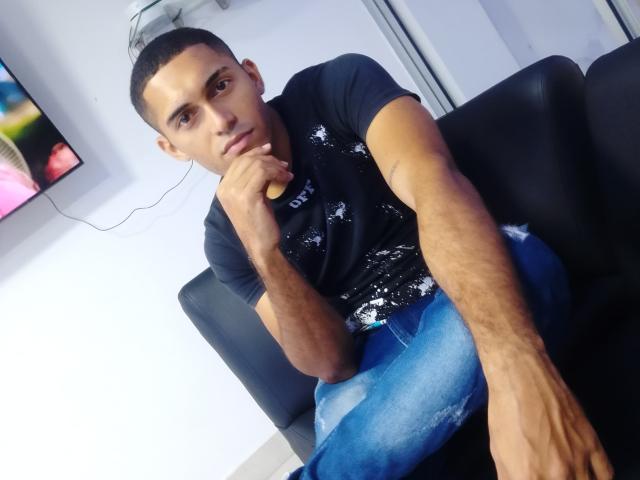 DereckStorm, with short hair and tan skin, sits on a black couch in a black t-shirt and blue jeans, resting his chin on his hand with a serious expression.