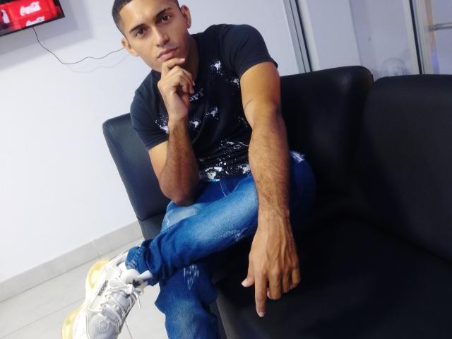 DereckStorm, sitting on a black couch, wearing a black t-shirt, blue jeans, and white sneakers, with a serious expression, resting his chin on his hand.
