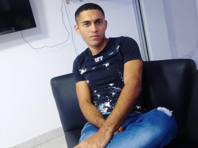 DereckStorm, short-haired, brown-skinned, wearing a dark blue, white splatter t-shirt and blue, ripped jeans, sitting on a black leather couch, looking slightly to the side.