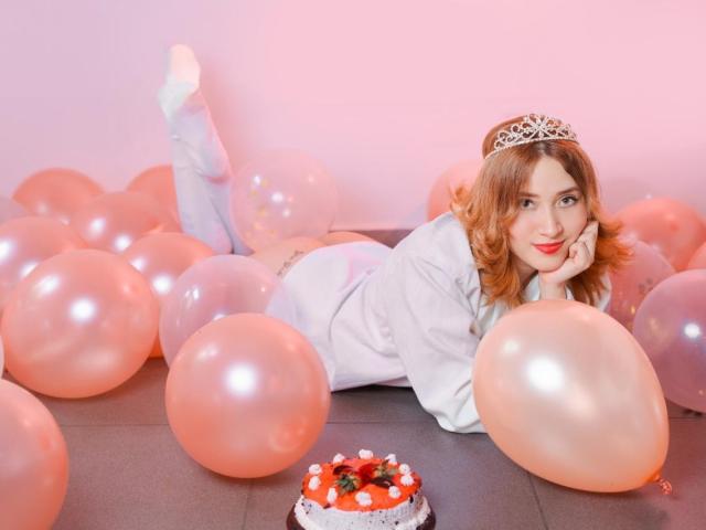 AnyHerrera with red hair, white socks, and a silver tiara, lying on pink balloons, wearing a white shirt, next to a small pink cake, smiling at the camera.