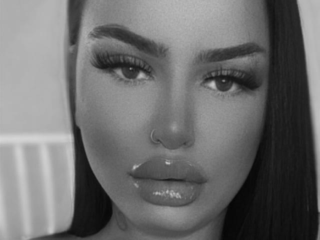 AriyaLux's face, black and white, showing glossy lips, nose ring, and full eyelashes, with long dark hair on the right, looking directly at the camera.