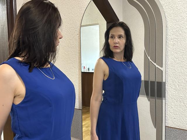 KarinaSwan with dark hair, blue sleeveless dress, small tits, standing in front of mirror, reflecting her serious face, wearing a silver necklace, in a room with white walls.