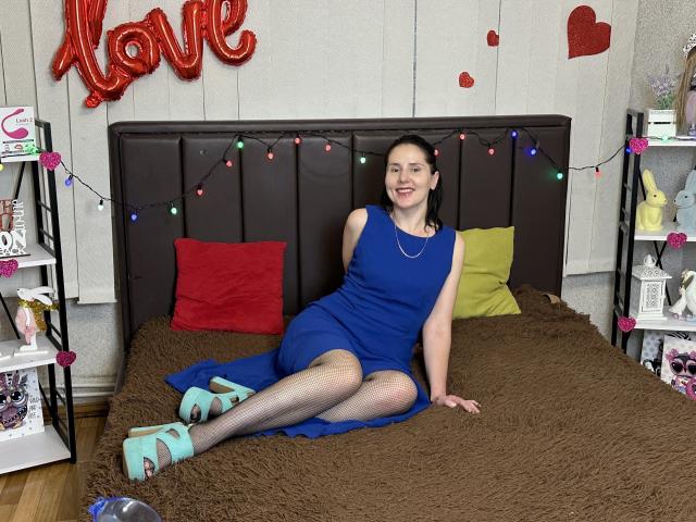 KarinaSwan with blue dress, fishnet stockings, and turquoise heels, sitting on brown bed, red and green pillows, black headboard, Christmas lights, and red heart decorations. Smiling, dark hair.