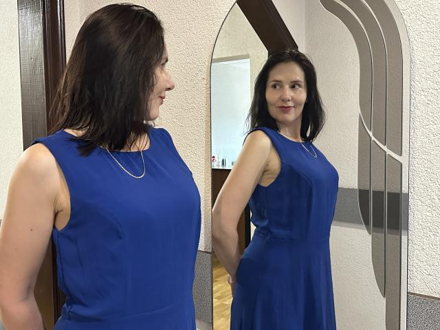 KarinaSwan with dark brown hair wearing a sleeveless blue dress, standing in front of a mirror showing her reflection. She has a slim build and is smiling.