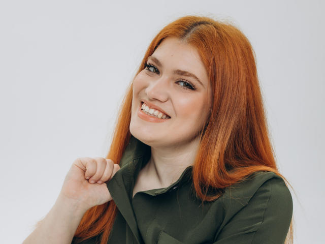 LindaSauver with long red hair, smiling, holding green shirt collar, fair skin, cute, and attractive, with subtle makeup, and natural teeth, looking slightly to the side.