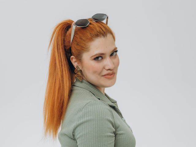 LindaSauver with orange ponytail, green ribbed shirt, and sunglasses on head, looking over shoulder with subtle smile, showing her ass and small tits, wearing gold earrings.